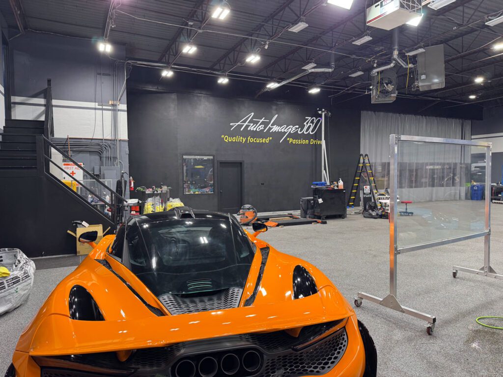 ArtWall Pro mural installation at Auto Image 360, featuring the company logo and slogan on a black wall with an orange sports car in the foreground.