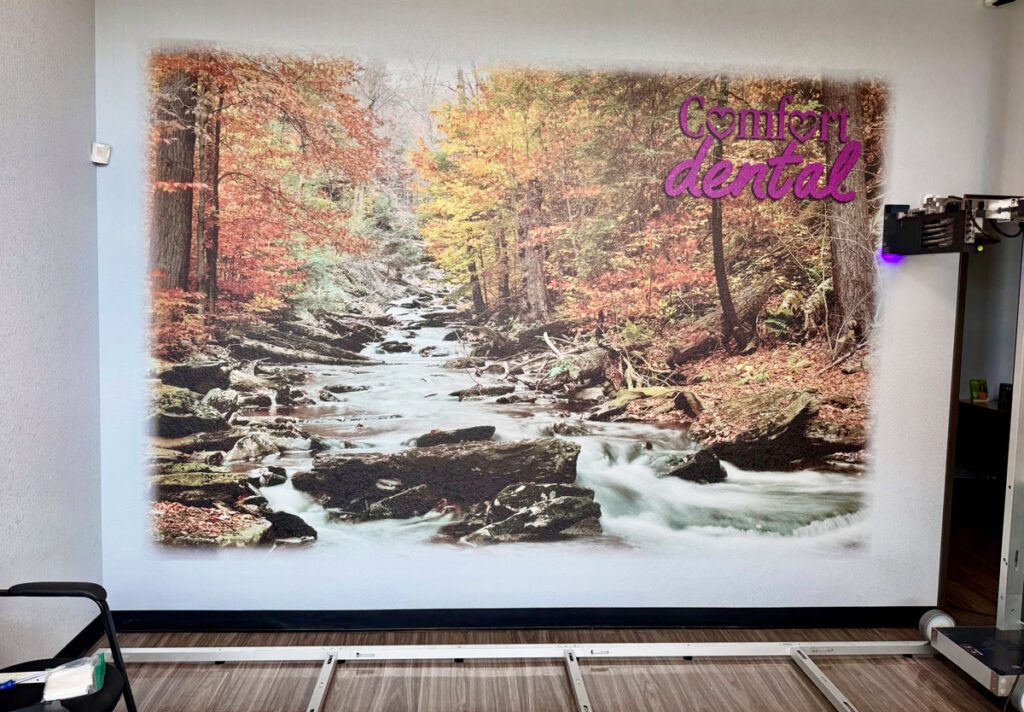 ArtWall Pro mural at Comfort Dental featuring a scenic forest stream with autumn foliage and the Comfort Dental logo printed above the design.